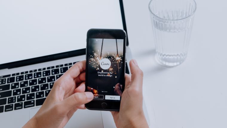 Hånd på iphone med canva, pc og glas vand i baggrund